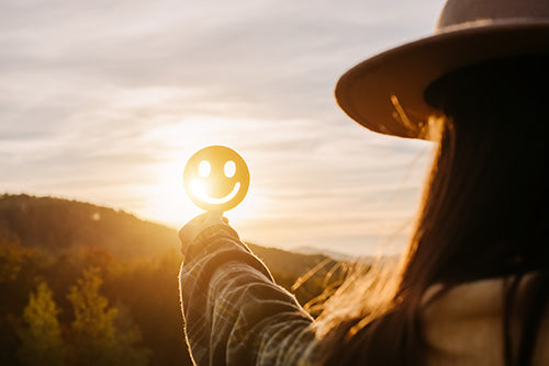 "Vrouw houdt een stralende smiley vast tegen de achtergrond van een zonsondergang, symboliserend de positieve energie en klanttevredenheid bij Vibali.